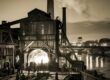 A gritty, sepia-toned illustration of a 19th-century Pittsburgh iron furnace roaring to life at night, workers silhouetted against the molten glow, with the Monongahela River in the background.