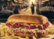 A nostalgic, warmly lit illustration of a Primanti Brothers sandwich — fries and slaw spilling out — set against a backdrop of Pittsburgh’s Strip District in the 1940s, with steelworkers and produce trucks in the scene.