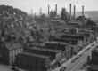 A rusting steel mill in the Mon Valley with smoke stacks silhouetted against a gray sky, symbolizing the industrial decline of towns like Braddock and Homestead.