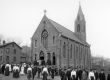 A black-and-white photograph of St. Anthony’s Chapel around 1900, showing its Gothic architecture, arched windows, and bell tower. People in period clothing gather outside after Mass, capturing the religious and community life of Troy Hill.