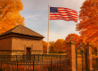 The Fort Pitt Block House in Pittsburgh on a sunny autumn day, with vibrant orange and red fall foliage, a tall American flag waving beside it, and an iron fence surrounding the historic brick structure.