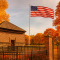 The Fort Pitt Block House in Pittsburgh on a sunny autumn day, with vibrant orange and red fall foliage, a tall American flag waving beside it, and an iron fence surrounding the historic brick structure.