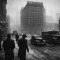 Black and white winter street scene in downtown Pittsburgh in the late 1940s with the Fort Pitt Hotel towering above Penn Avenue, vintage cars and pedestrians in coats and fedoras, and light snow in the air.