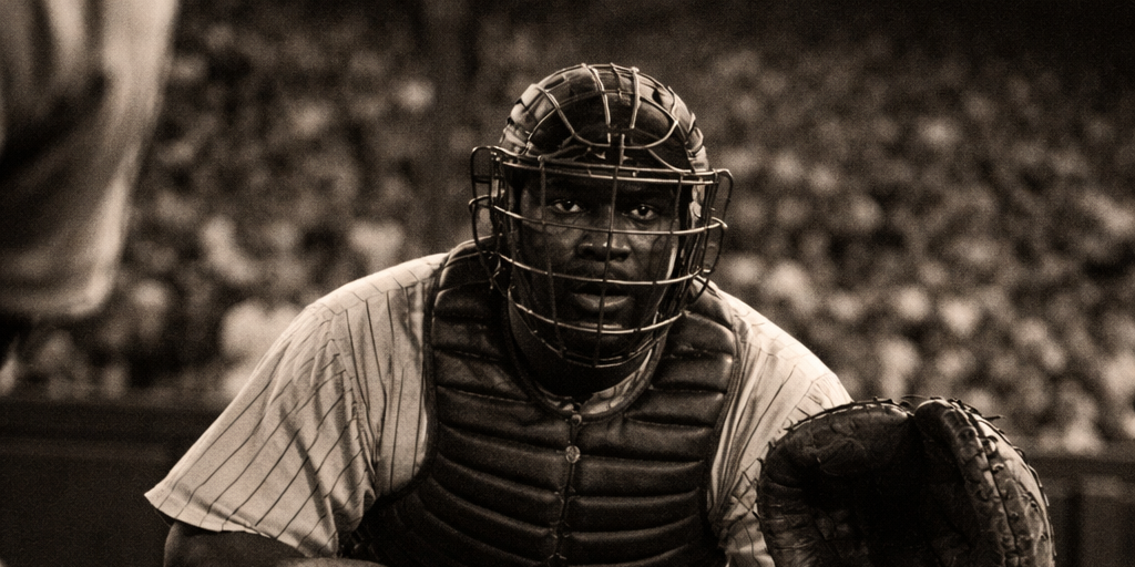 Baseball catcher in 1940s uniform crouches behind home plate at a packed Pittsburgh stadium, capturing the era when Josh Gibson was considered the greatest hitter in baseball.