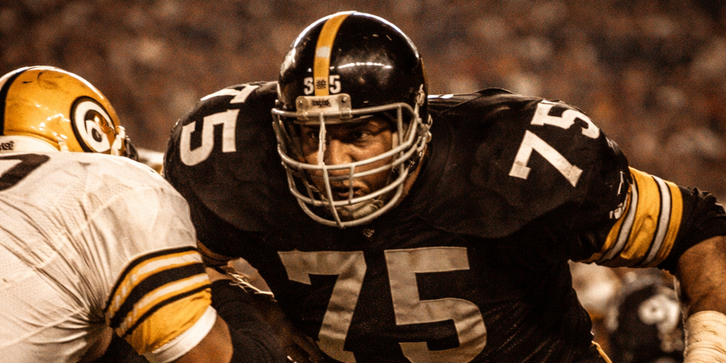 A Pittsburgh Steelers defensive lineman in the classic 1970s black and gold uniform breaks through an offensive line under stadium lights, capturing the intensity of the Steel Curtain defense.